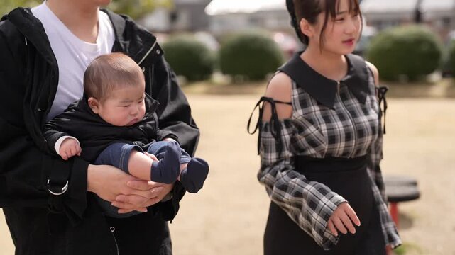 Man holds baby gently in park setting. Child smiles softly, eyes wide with wonder. Bright yellow tent stands behind bare trees. Distant pine tree adds depth to natural backdrop. Sunlight warms scene