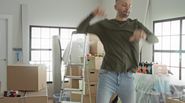 Young man dancing with cardboard boxes, arms raised amid packed moving boxes in a bright building; fresh start joy.