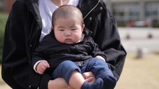 Man holds baby gently in park setting. Child smiles softly, eyes wide with wonder. Bright yellow tent stands behind bare trees. Distant pine tree adds depth to natural backdrop. Sunlight warms scene