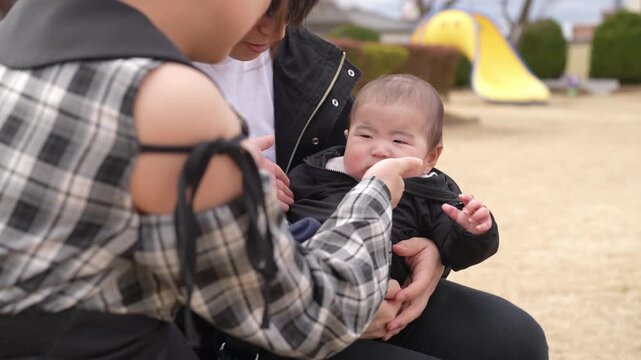 Man holds baby gently in park setting. Child smiles softly, eyes wide with wonder. Bright yellow tent stands behind bare trees. Distant pine tree adds depth to natural backdrop. Sunlight warms scene