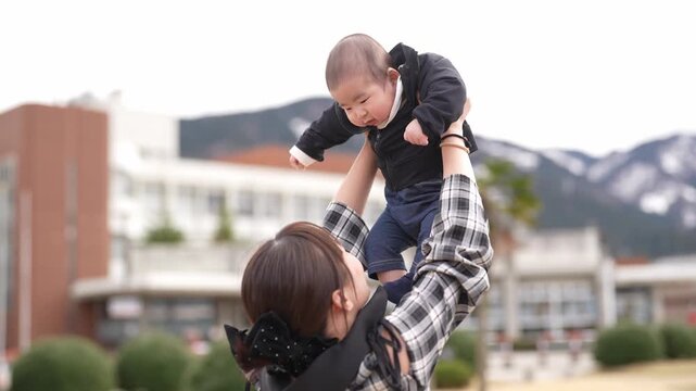 Man holds baby gently in park setting. Child smiles softly, eyes wide with wonder. Bright yellow tent stands behind bare trees. Distant pine tree adds depth to natural backdrop. Sunlight warms scene