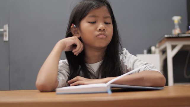 Bored little Asian girl leaning on hand while reading a thick book, tired child feeling unmotivated or sleepy during homework, academic boredom and learning difficulties
