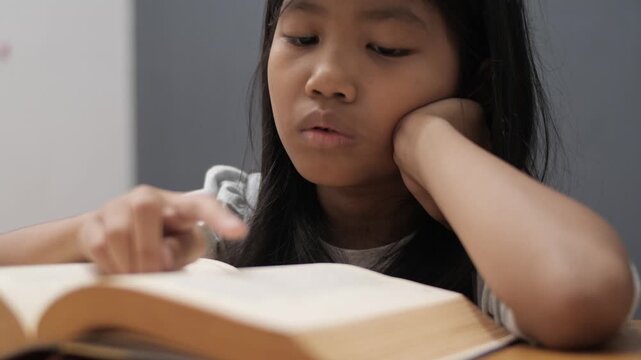Bored little Asian girl leaning on hand while reading a thick book, tired child feeling unmotivated or sleepy during homework, academic boredom and learning difficulties