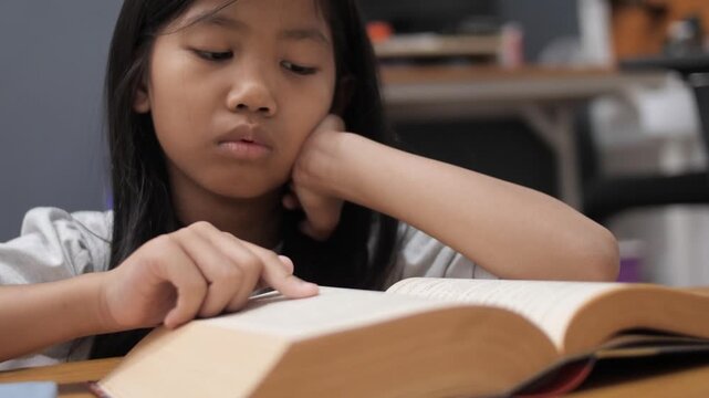 Bored little Asian girl leaning on hand while reading a thick book, tired child feeling unmotivated or sleepy during homework, academic boredom and learning difficulties