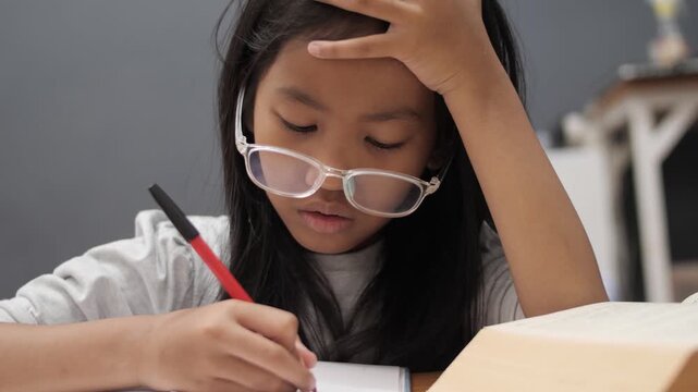 Stressed little Asian girl with glasses crying while studying, unhappy child having headache or difficulty with homework, academic pressure and learning disability