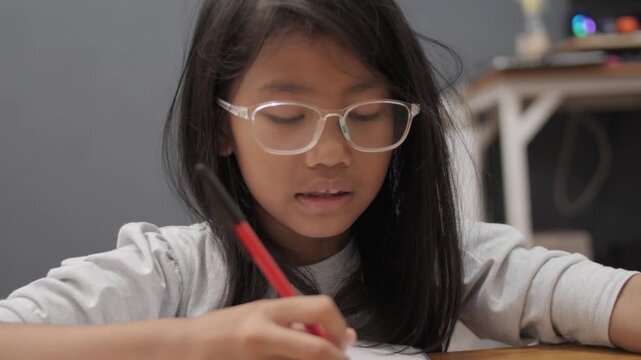 Diligent little Asian girl with glasses writing in notebook, focused child studying and reading book at home, early education and homeschooling
