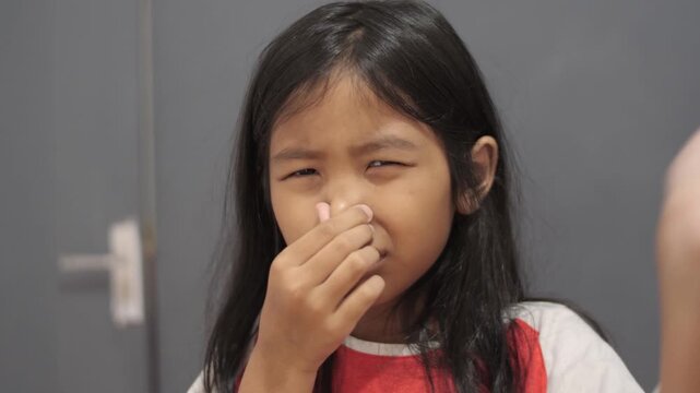 Disgusted little Asian girl pinching her nose due to bad smell, child reacting to unpleasant odor or stinky scent with funny expression, air pollution