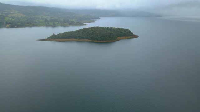 A serene aerial view of Cocha Lagoon showcasing a small island surrounded by still water and lush green hills.