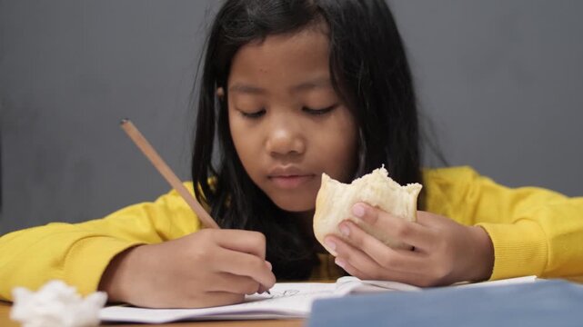 Focused little Asian girl drawing in notebook while eating a snack, tired child multitasking with homework and food, early education and childhood lifestyle