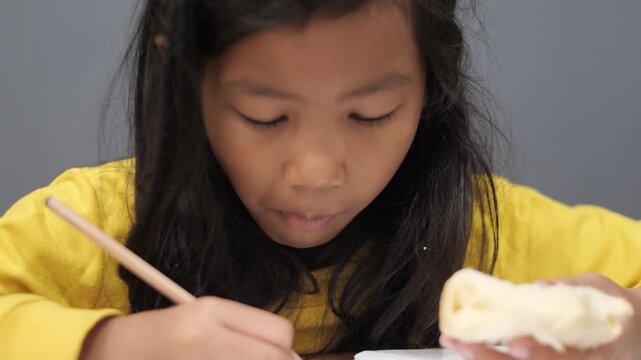 Focused little Asian girl drawing in notebook while eating a snack, tired child multitasking with homework and food, early education and childhood lifestyle