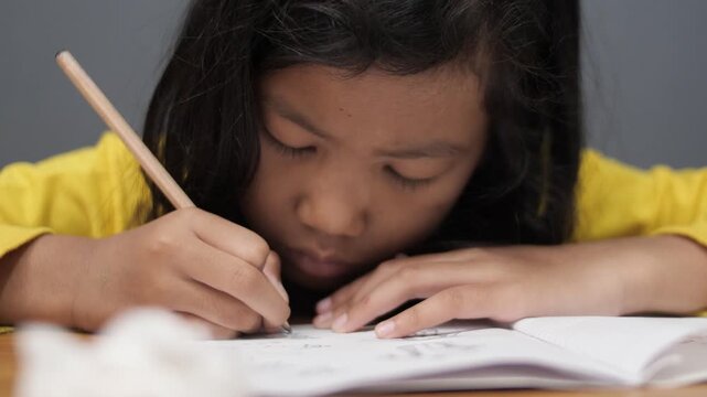 Diligent Asian little girl in yellow sweater writing on her notebook. Close up of focused elementary student doing homework or drawing. Childhood education, homeschooling
