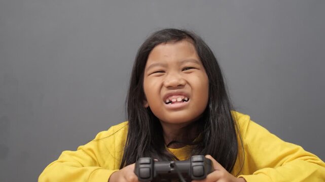 Frustrated little Asian girl grimacing while playing video games, upset child losing a game holding controller, gaming challenge and emotional reaction