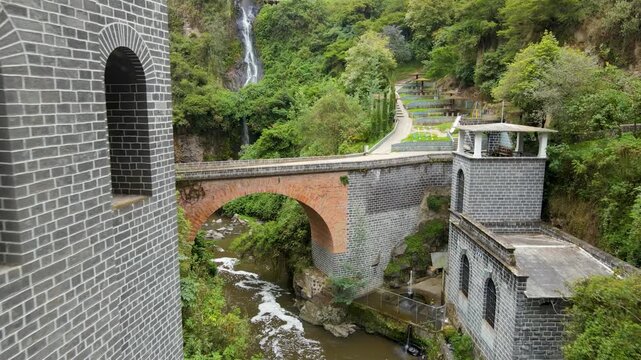 Camera moves from a gray stone church across a red-arched bridge toward a tall cascading waterfall and terraced path on the green cliffside.