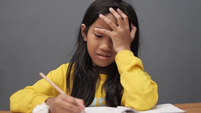Stressed little Asian girl with glasses crying while studying, unhappy child having headache or difficulty with homework, academic pressure and learning disability
