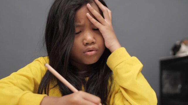 Stressed little Asian girl with glasses crying while studying, unhappy child having headache or difficulty with homework, academic pressure and learning disability