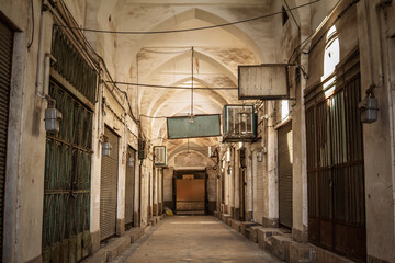 Fototapeta premium An empty covered passage in Yazd Khan Bazaar, Iran, with vaulted arches, hanging signs, and closed shop shutters in afternoon light, conveying a quiet, deserted moment in a historic marketplace.