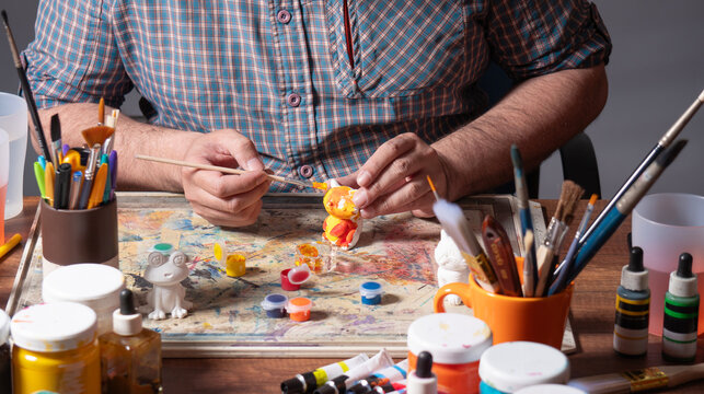 A man's hands in a blue shirt holding a fine brush with orange paint are painting a small white ceramic figure