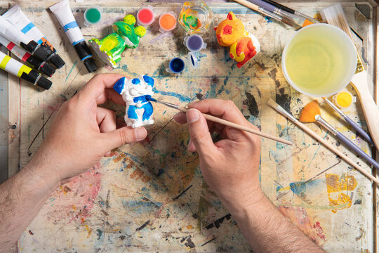 man's hands in a blue shirt holding a fine brush with orange paint, painting a small white ceramic figure on a work table