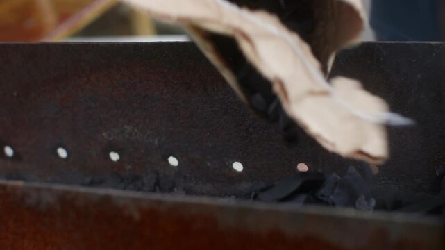 Closeup hand placing paper on charcoal, home cook prepping backyard grill with crumpled newspaper fed into rusty metal basin over briquettes, textured ash and ember hints, soft evening