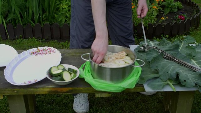 Caucasian man seasoning potato slices outdoors, stainless pot and bowl of zucchini on wooden bench, floral border in background, hands sprinkling spice over sliced tubers, prepping