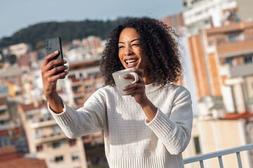 Fototapeta premium Beautiful woman drinking a cup of tea while doing a videocall on her balcony at home
