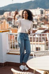 Fototapeta premium Beautiful woman drinking a cup of tea while doing a videocall on her balcony at home