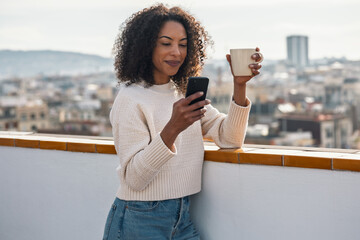 Fototapeta premium Beautiful woman drinking a cup of tea while doing a videocall on her balcony at home