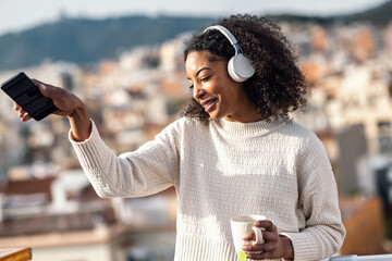Fototapeta premium Stylish woman drinking a cup of tea while dancing and listening to music with headphones on her balcony at home