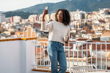 Fototapeta premium Beautiful woman drinking a cup of tea while doing a videocall on her balcony at home
