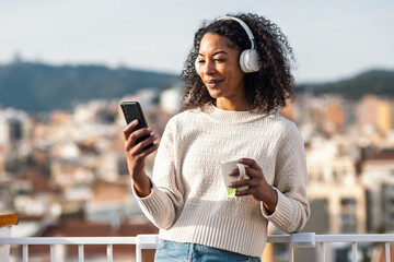 Fototapeta premium Pretty woman drinking a cup of tea while using smartphone with headphones on her balcony at home
