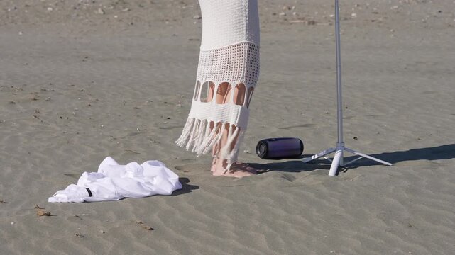Young social media influencer preparing for a photoshoot on a sunny beach, taking off her white shirt and setting up her phone on a selfie stick