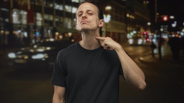 Man young hispanic with long hair points finger to neck on a city street at night, wearing black t shirt and stud earring; defiance.