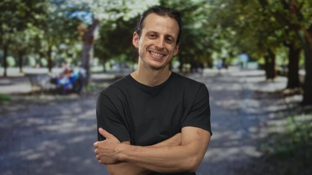 Man smiling with arms crossed showing forearms on a sunlit park pathway under green trees and dappled shade; confidence ease.
