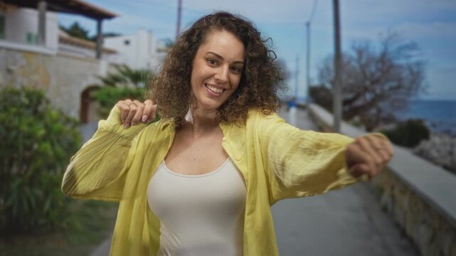 Woman smiling with raised arms and loose yellow shirt revealing cleavage on seaside street promenade; joy celebration.