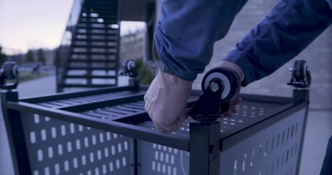 Man screwing wheel to upside down metal cart at dusk - medium shot