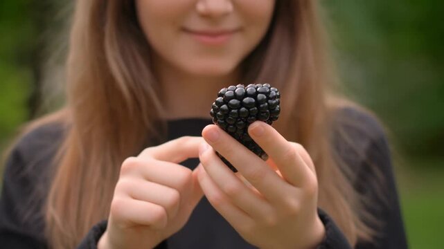 A young person delicately holds a fresh, dark, aggregate fruit. Their expression suggests a pleasant emotion with flowing brown hair, and blurred green background