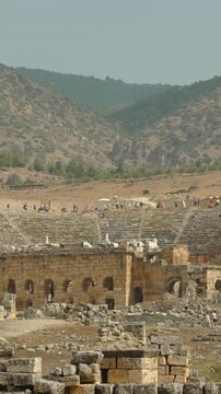 Vertical video. Ancient Ruined Amphitheater in the Distance at the Foot of the Mountain, People on Top of It.