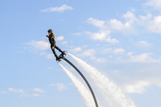 A flyboarder rises high above the sparkling water, propelled by powerful jets of water. The individual balances skillfully on the board, wearing a helmet and wetsuit, showcasing a daring pose.