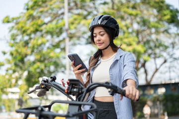 Naklejka premium Pretty asian woman wearing helmet looking at smartphone while riding e bike electric bicycle in park