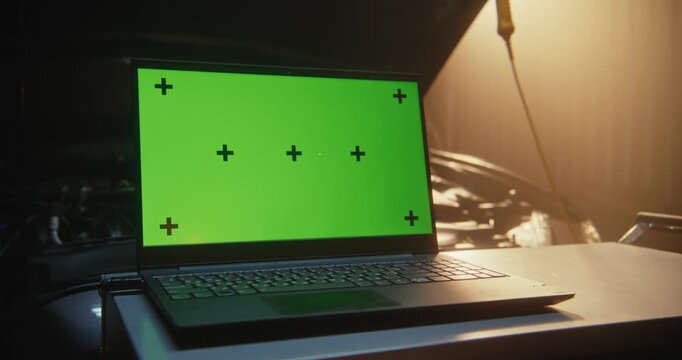 Laptop With Green Screen and Black Tracking Points, Ideal For Motion Tracking and Screen Compositing, Positioned Next to Open Car Hood in Sun Drenched Workshop. Post Production and Digital Marketing.