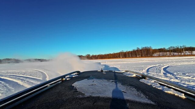 Roof-mounted view of a car drifting through snow in bright sunshine under a clear blue winter sky.