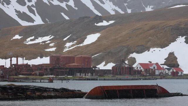 Snowy view of Grytviken whaling station ruins across a cold harbor in South Georgia, with rusted tanks and mountains&mdash;remote polar history and industrial decay.