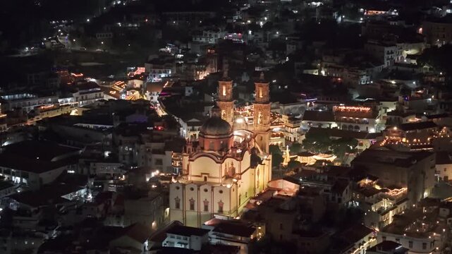 Telephoto aerial view of the Santa Prisca temple at night, viceroyalty architecture with illuminated and interconnected streets of Taxco, Guerrero, Mexico