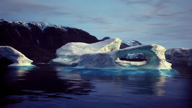 Sculpted ice arch in Greenland seascape with carved openings and pale turquoise core, reflective calm water, textured surface and distant dark headlands, quiet