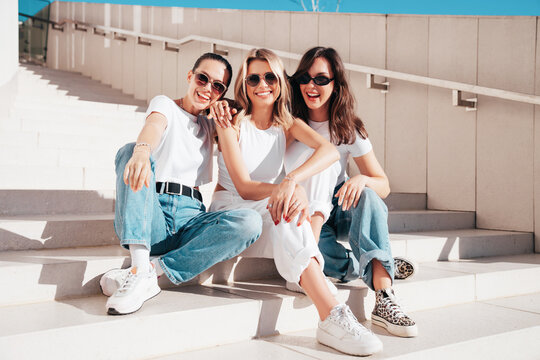 Three young beautiful smiling hipster female in trendy summer t-shirt  and jeans clothes. Sexy carefree women posing in street. Positive models having fun, sit at stairs. In sunny day, sunglasses