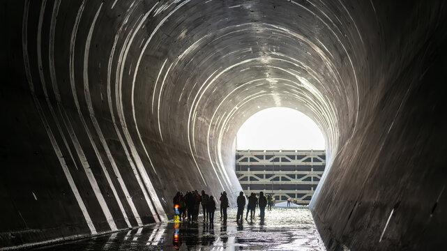 Silhouette of workers inside a large concrete tunnel at a dam construction site, with natural light at the tunnel exit.