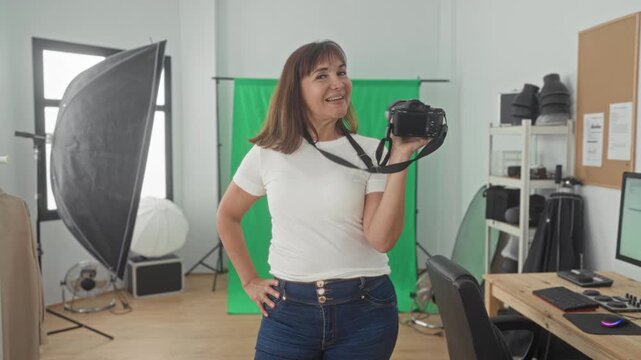 Woman holding camera with hand on hip in studio with green screen, softbox lighting, computer desk and monitor visible; confidence creativity.