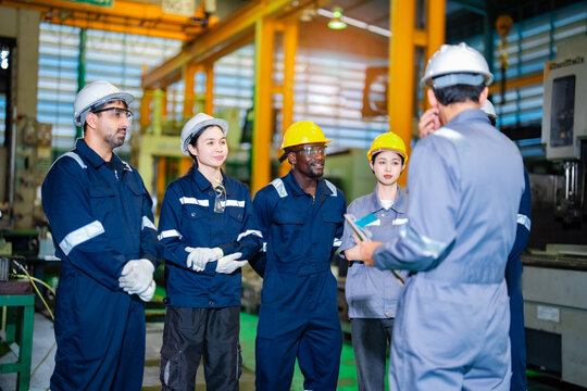 Industrial workers group standing with supervisor during briefing in manufacturing workshop with safety gear and helmets. Team focused on communication. Concept of leadership, operational efficiency.