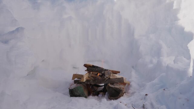 Small campfire burning inside a deep snow pit in winter. Thin smoke rises above the stacked firewood while flames slowly burn in the cold snowy environment. High quality 4k footage