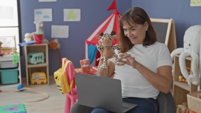 Woman holding a giraffe puppet and waving to laptop in a kindergarten building; online teaching joy.